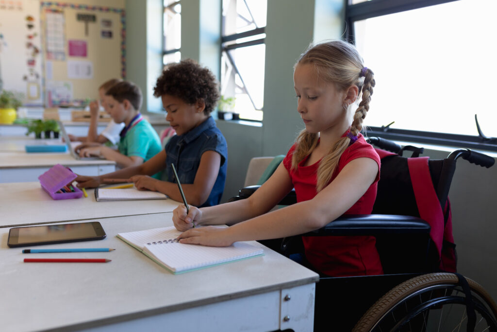 Side view of a Caucasian schoolgirl with blonde hair in plaits sitting in a wheelchair and sitting at a desk writing, with a diverse group of schoolchildren sitting at desks beside her working in an elementary school classroom